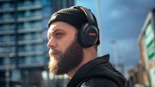 A bearded man in an urban area on a sunny day wearing a beanie and a set of Orange 'O' Edition Mk II ANC headphones