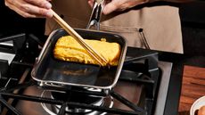 A square omlete pan on a gass stove, someone using a pair of chopsticks to roll it.