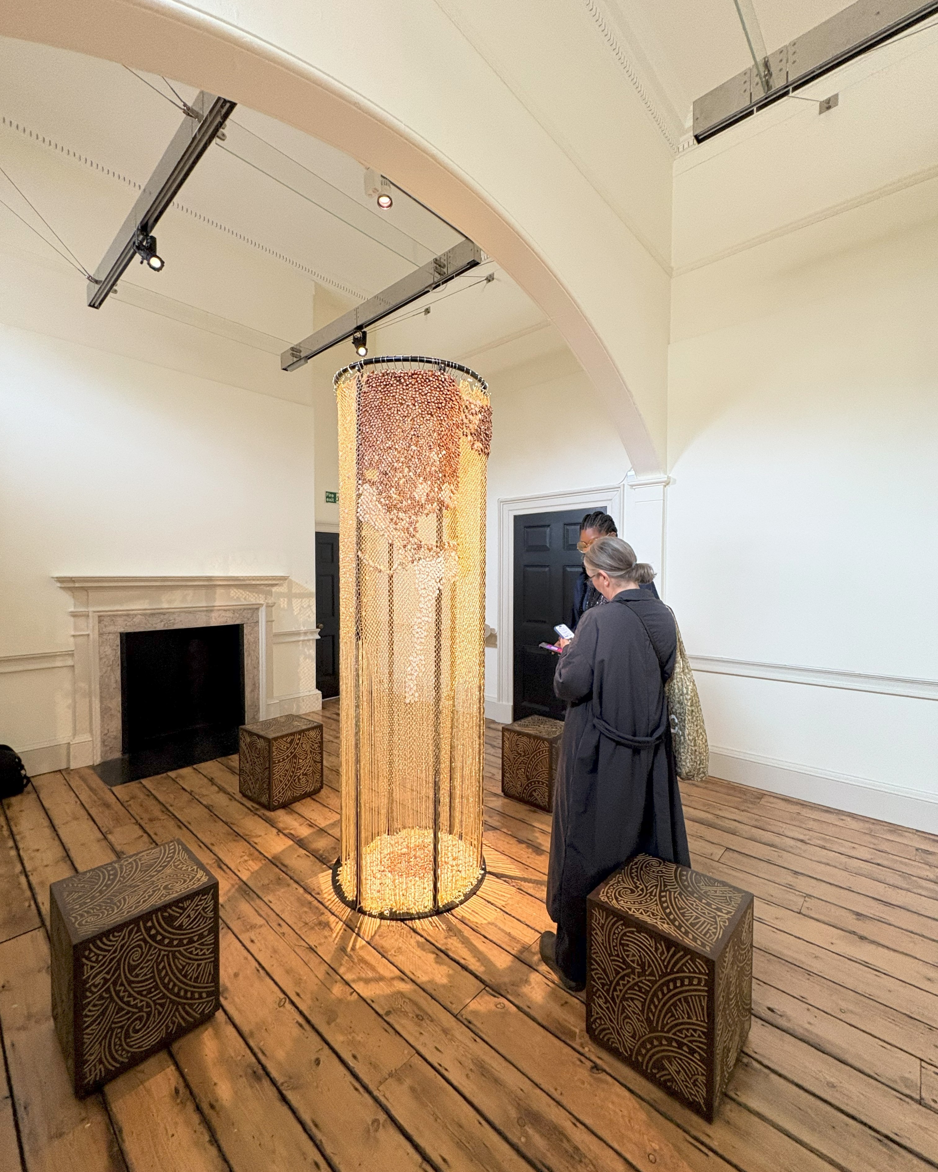 A standing installation made of woven metallic fibers and shells sits next to four square, textural stools decorated in abstract patterns in a wood-clad gallery as two women stand next to it.