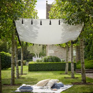 White sail shade with black tassels strung between a line of trees on a lawn, with a picnic blanket and cushions beneath and a stone building in the background