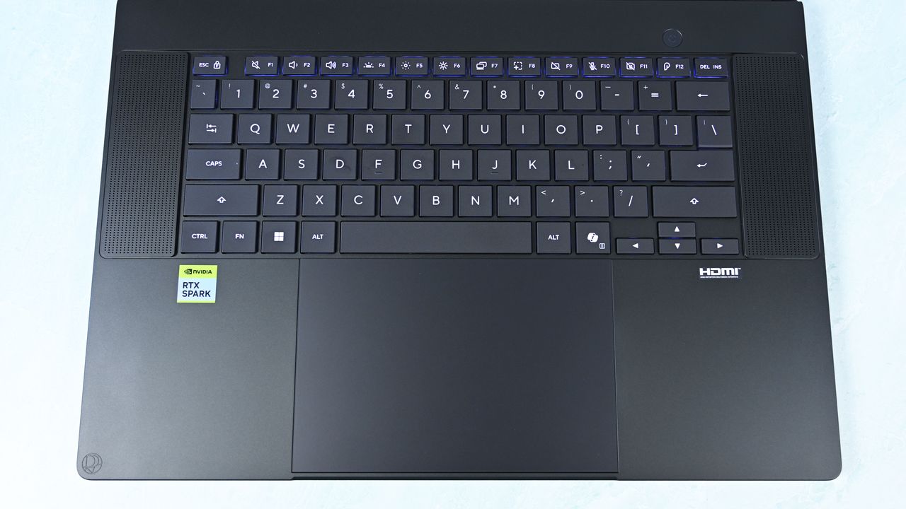 Top-down view of the keyboard deck showing flanking speaker grilles, power button, trackpad with DialPad icon, and RTX Spark sticker.