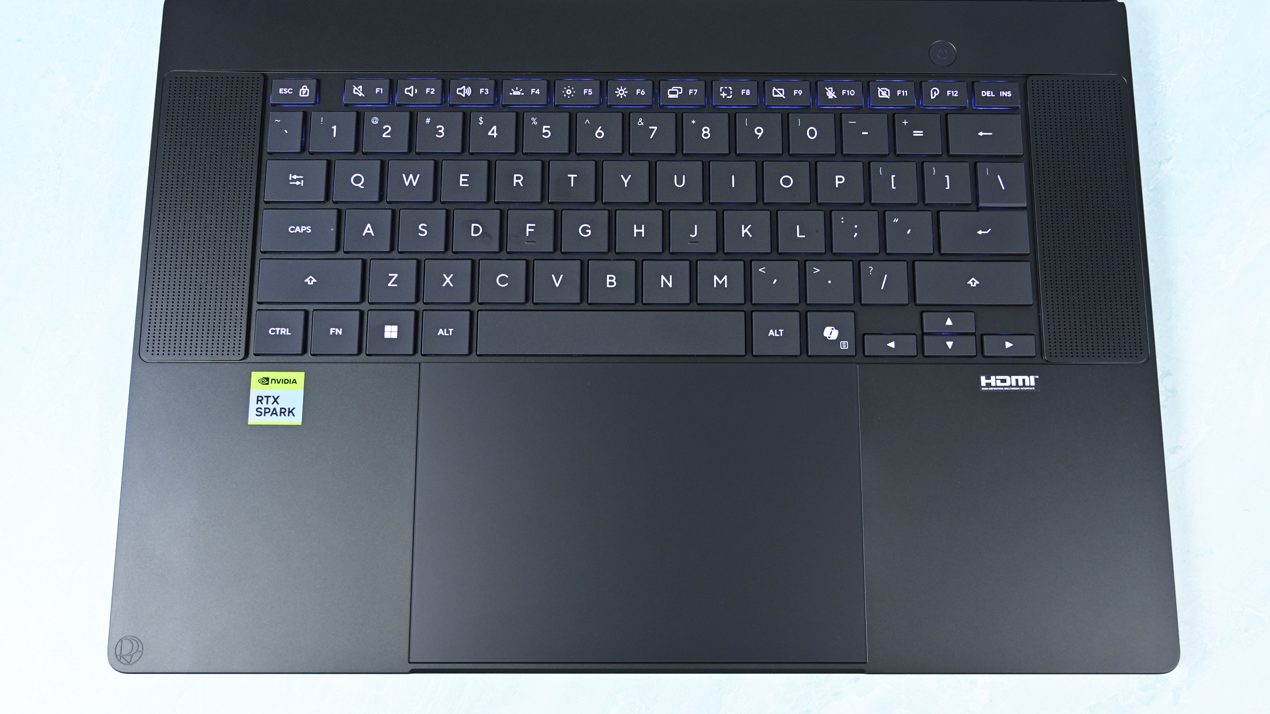 Top-down view of the keyboard deck showing flanking speaker grilles, power button, trackpad with DialPad icon, and RTX Spark sticker.
