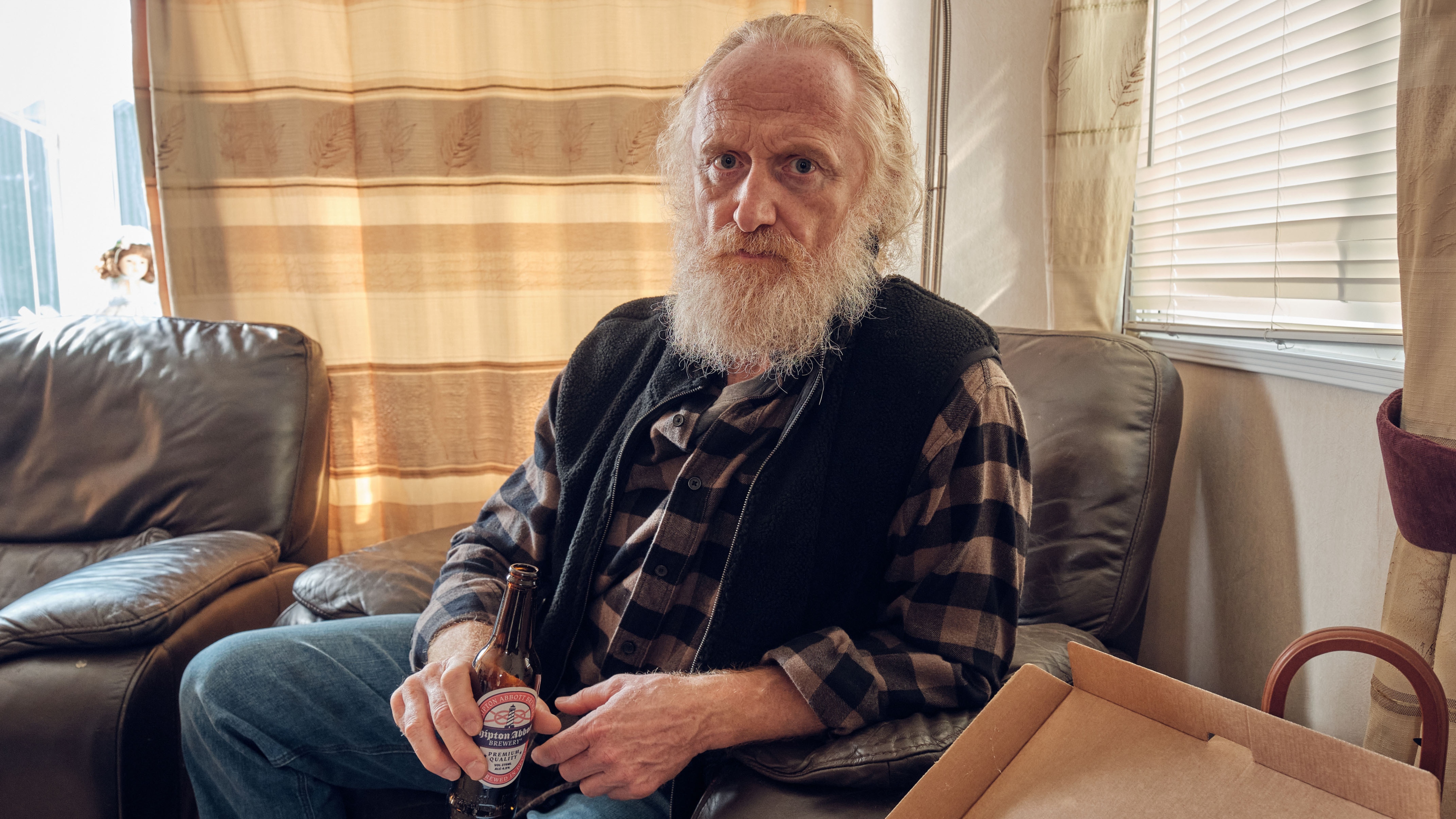 Trent Morgan (Murray McArthur) sits on a leather armchair in his static caravan. He has a white, whiskery beard that goes all the way round to his hairline, and his hair is receding at the front but longish at the back. He is holding an open bottle of Beer and looking into the camera.