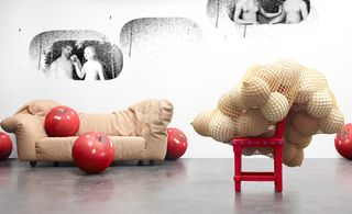 Brown sofa against with large sized red apples photographed against a white wall with painted on black and white wall art. On the right of the wall art is a net bag filled with balls and placed on a red chair