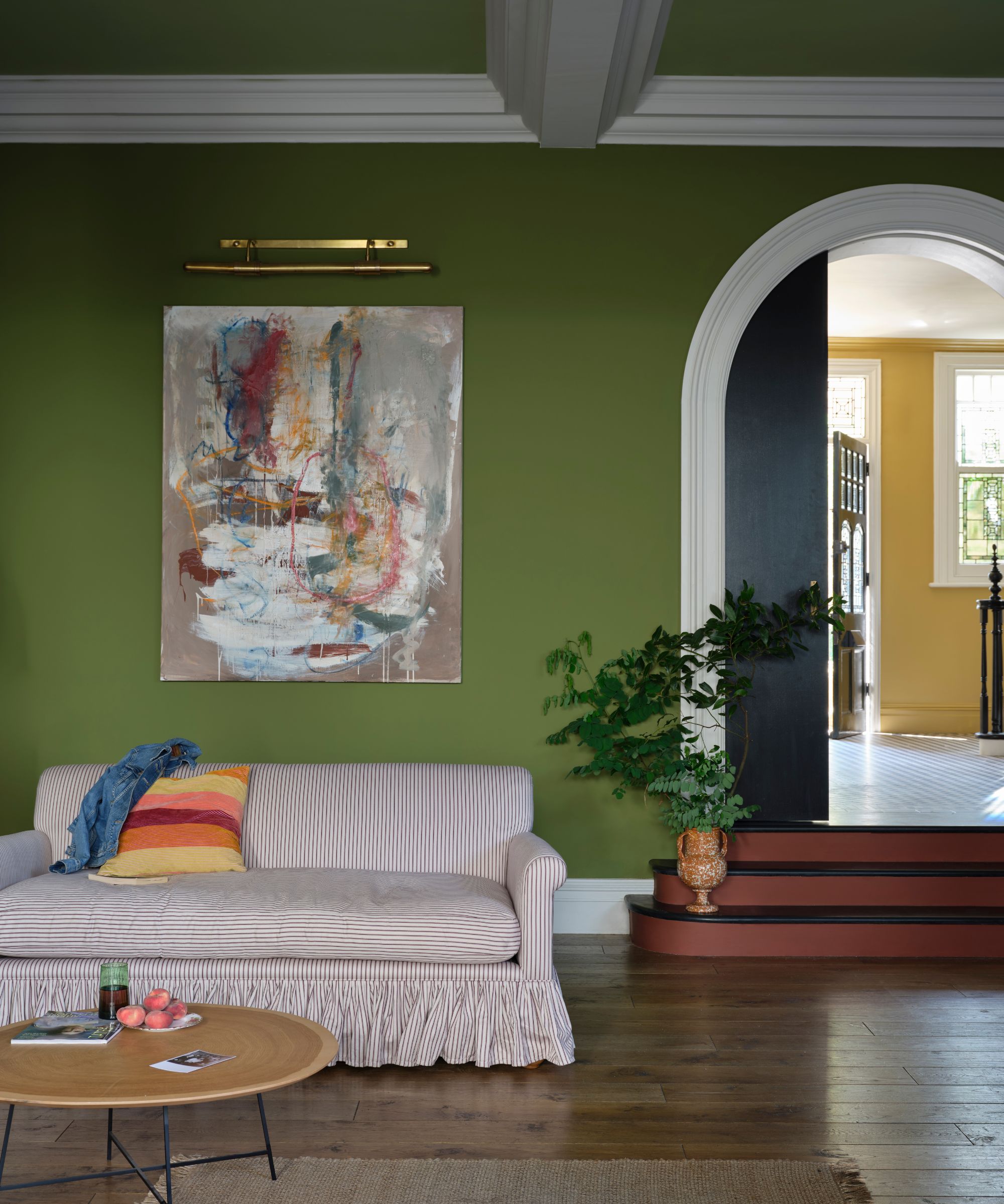 A cozy, low-light living room with moss green walls with white trim, abstract artwork with a picture light, a striped ruffle sofa, wood flooring, and an arched doorway.