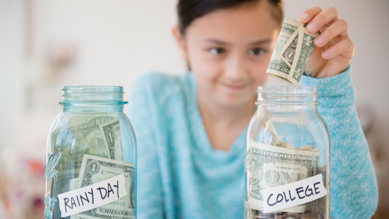 A young girl puts a dollar bill into a jar of bills and coins with a label that reads &amp;quot;College.&amp;quot;