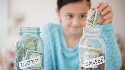 A young girl puts a dollar bill into a jar of bills and coins with a label that reads "College."