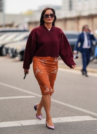 A guest wears dark brown sunglasses, dark red maroon oversized pullover, white t-shirt, silver earrings, dark burnt orange with silver detail over the knee skirt, shiny light pink heels, outside Prada, during the Milan Fashion Week Spring/Summer 2024-2025 on September 19, 2024 in Milan, Ital