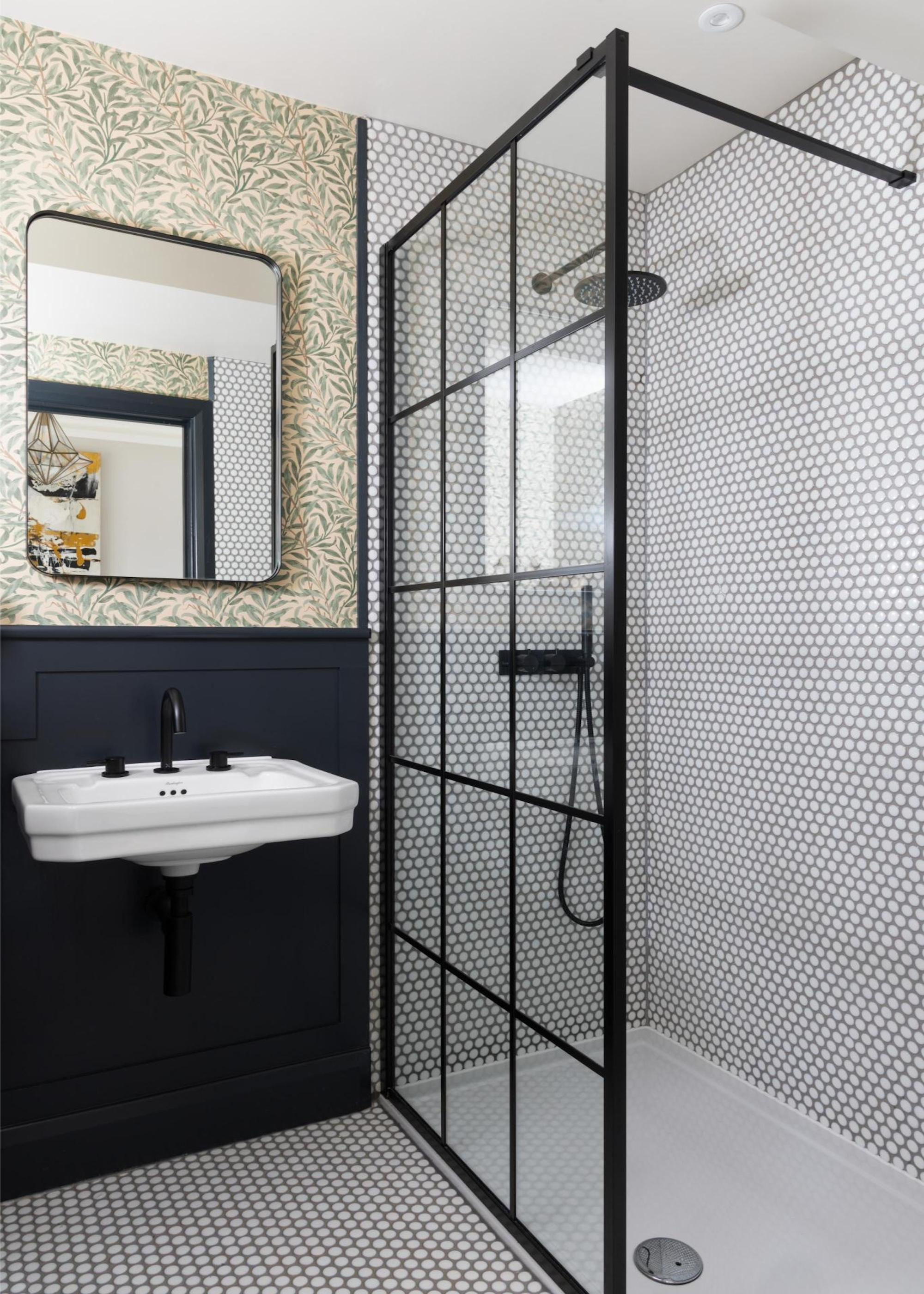 A small bathroom with a walk-in shower with a black-framed glass panel, mosaic tiles on the shower walls and flooring, and a patterned wallpaper above the floating sink and black-painted wall panel