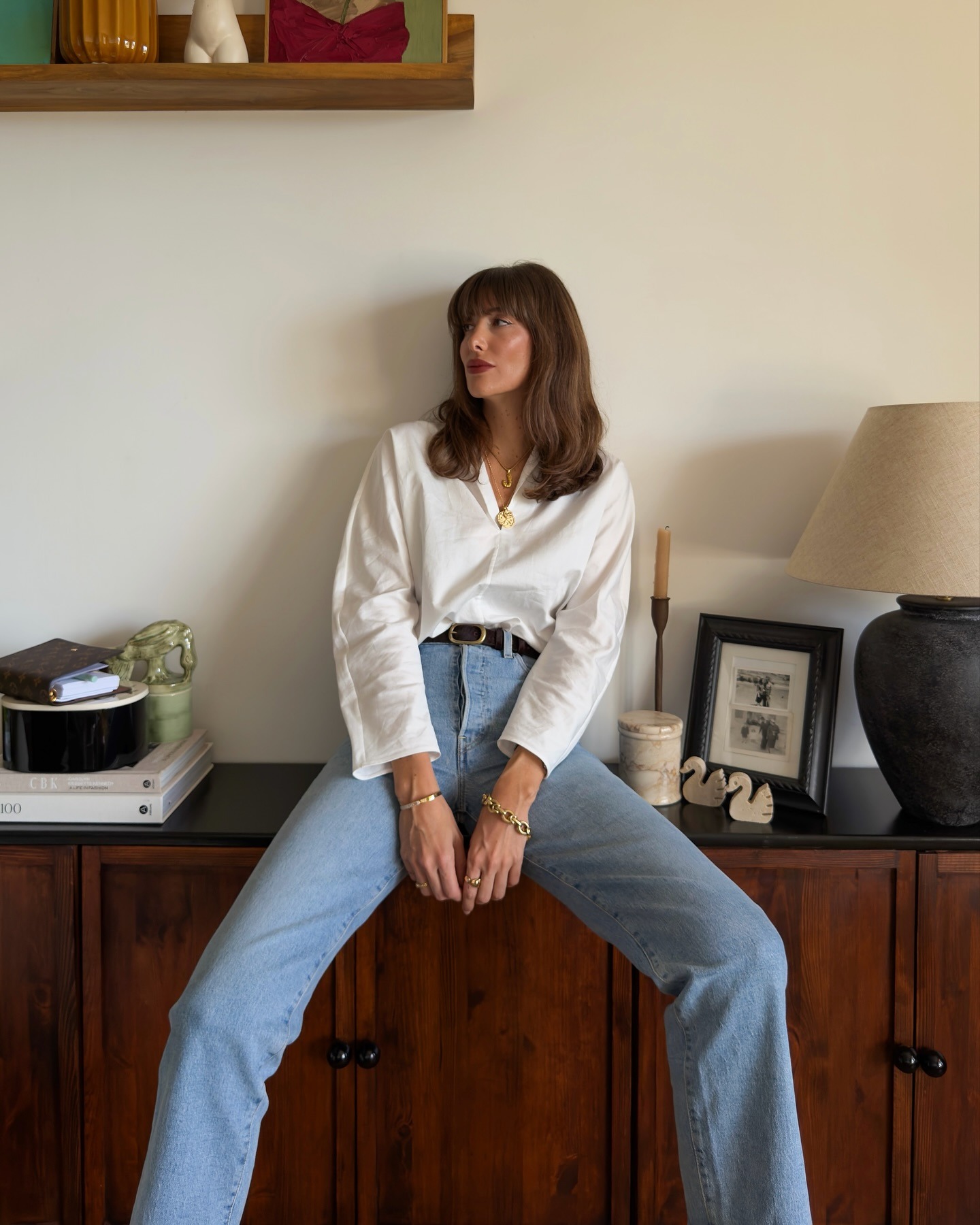 A woman leaning against a sideboard in her apartment, wearing blue jeans and a white shirt with a lob hairstyle