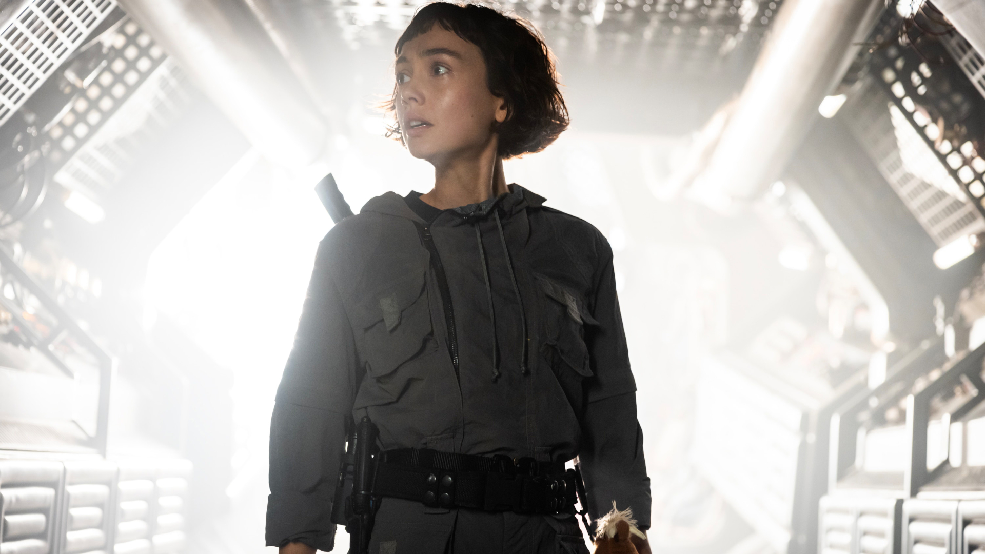 Still from the TV show Alien: Earth. A woman wearing a grey jumpsuit and a utility belt is standing in a bright tunnel. She is looking around, searching for something.