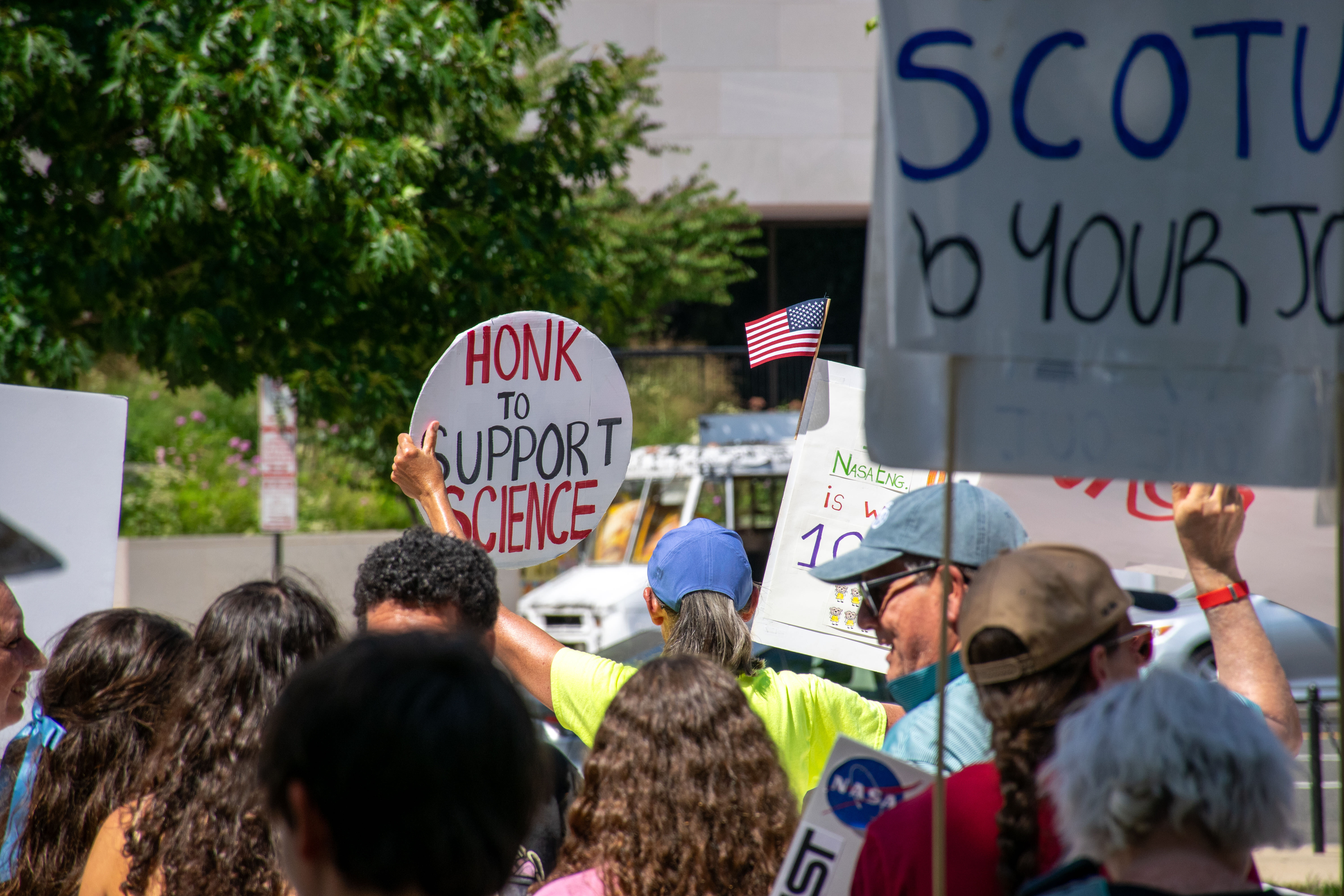 People hold signs outside on a sunny day in protest of NASA budget cuts, July 20, 2025.