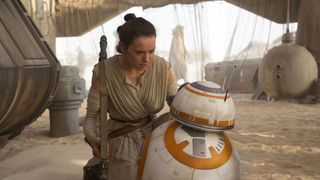 a woman bends down to talk to a small orange and white robot made up of two spherical parts.