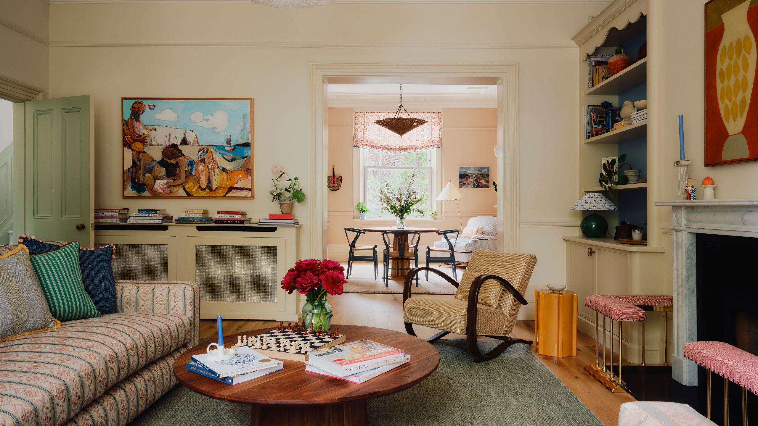 A large living room with cream walls, crown moulding, a large marble fireplace with a pink hearth, a low, round wooden coffee table, a mid-century armchair, and blue-painted shelving alcoves.
