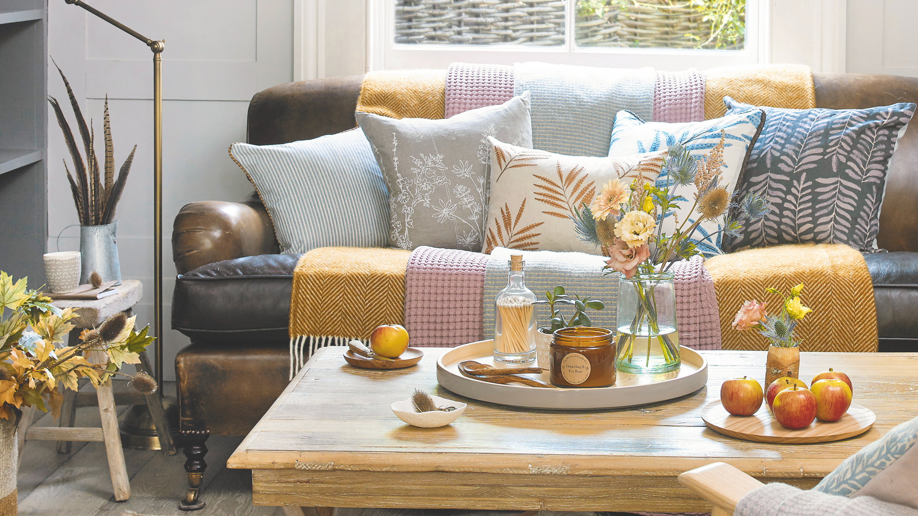 Living room with a traditional leather sofa and throws and cushions. Rug and brass floor light, and a tray and apples on a small table.