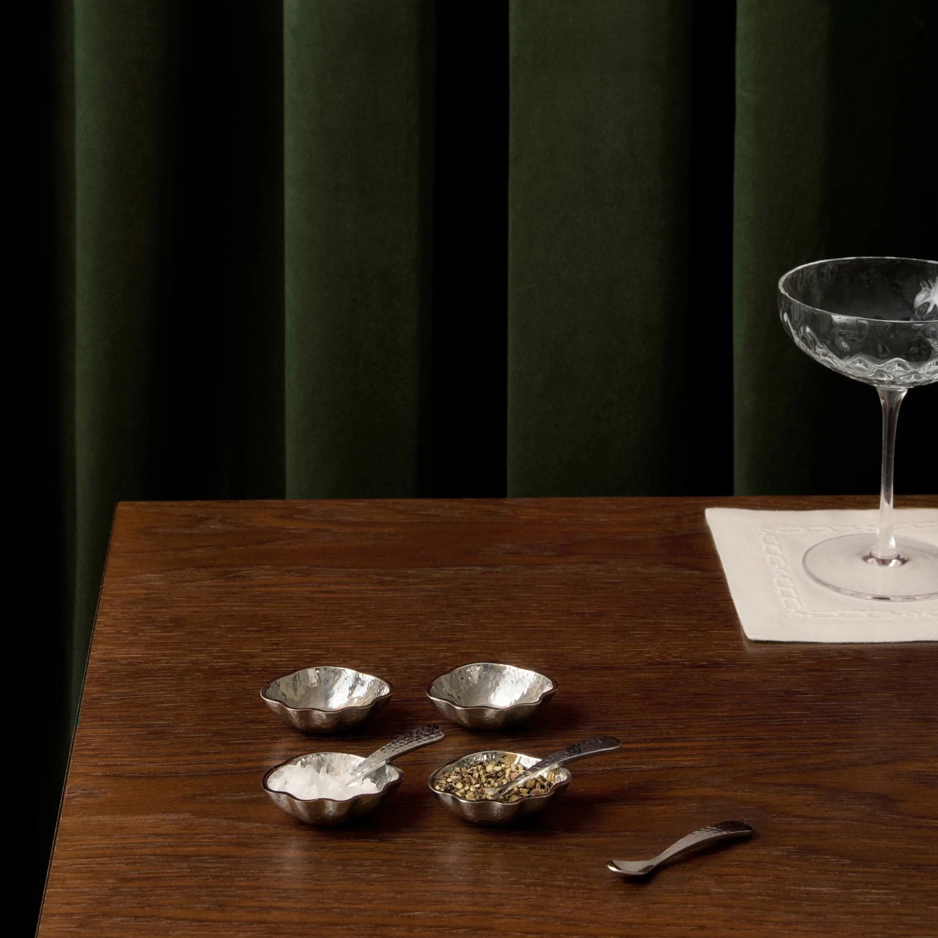 The Isla Salt & Pepper Shells with Spoons from Victoria Beckham's tableware collection with Westwing on a dark wood table with a green velvet curtain in the background