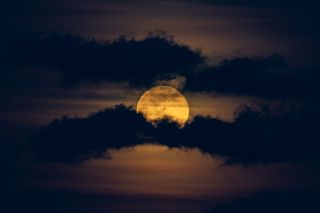 A bright yellow full moon shines behind two bands of dark clouds that streak across the foreground of the image.