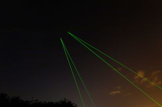Night image, dark cloudy sky, two sets of green laser beams projected up to the sky from ground level, of the space waste lab tracking system, silhouette of tree tops