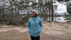 A woman on a beach near a lake wearing the Rab Microlight Alpine Women's Down Jacket with her hands in her pockets
