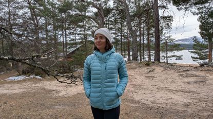 A woman on a beach near a lake wearing the Rab Microlight Alpine Women's Down Jacket with her hands in her pockets