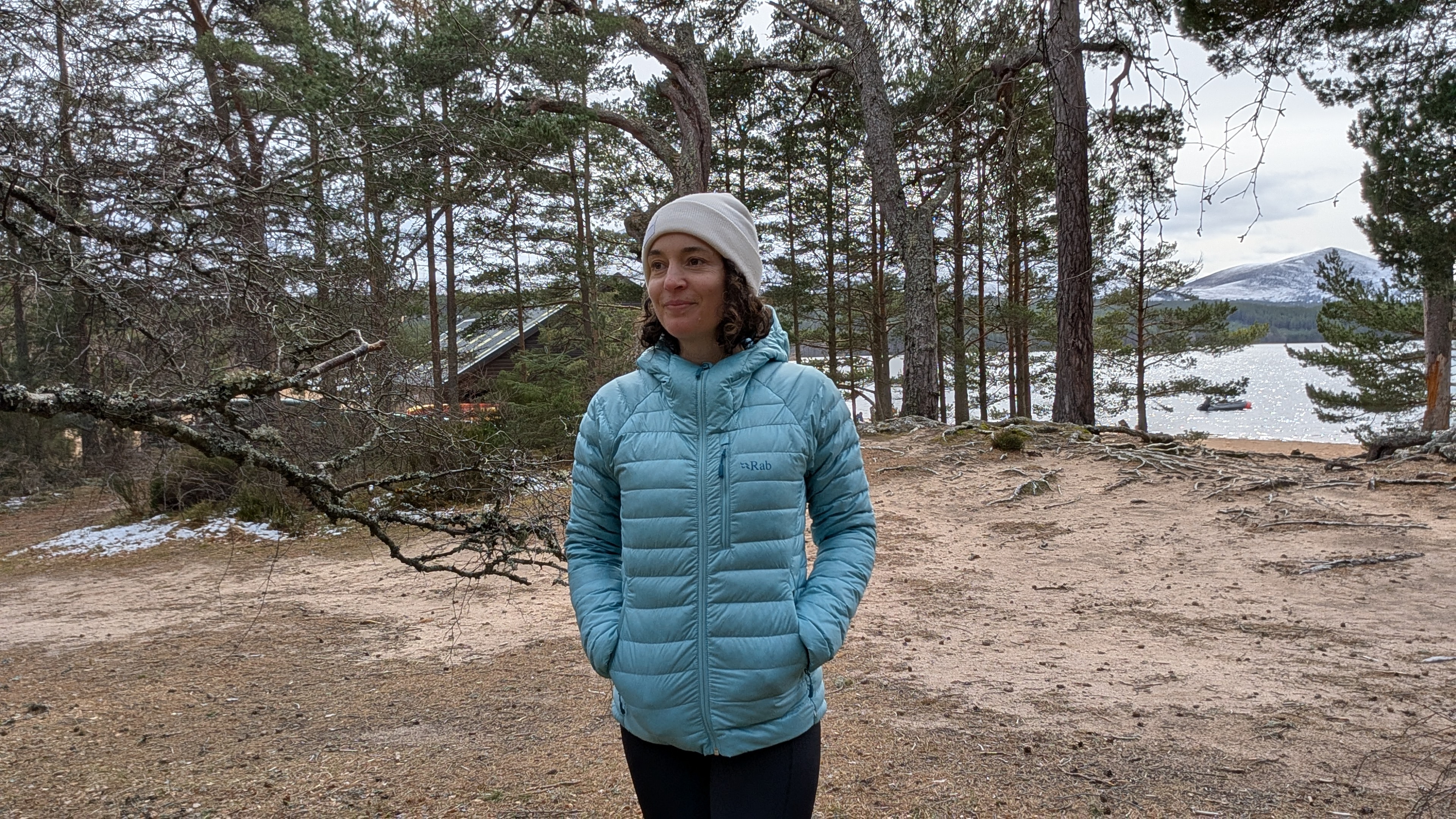 A woman on a beach near a lake wearing the Rab Microlight Alpine Women's Down Jacket with her hands in her pockets