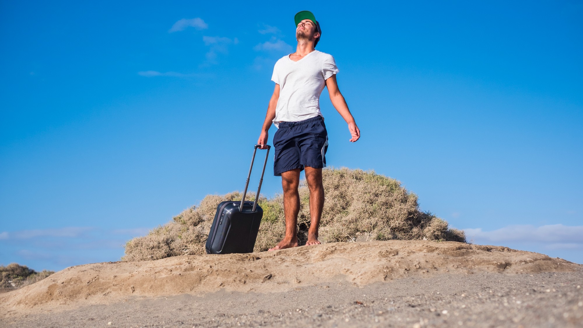 Young man standing with a carry-on suitcase on a beach 