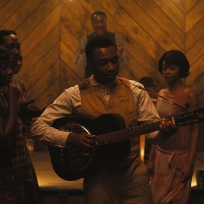 A man (Miles Caton as Sammie Moore) plays the guitar while standing in a crowd in a wooden barn, in 'Sinners.'