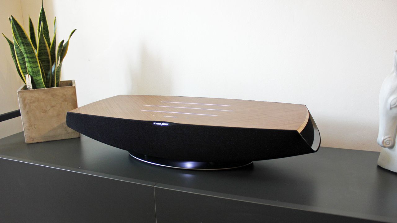 The Sonus Faber Omnia wireless speaker sitting on a black side table, next to a plant. We can see its boat-like shape, with a flat top surface and a curved body underneath. The flat top is wood, while the bottom is black.