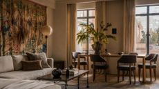 A cozy, warm living room with beige interiors, a minimalist color palette featuring brown chairs, a brown coffee table, a beige couch, a large wall tapestry, a standing lantern floor lamp, a dramatic vase of feathered stems and semi sheer window treatments