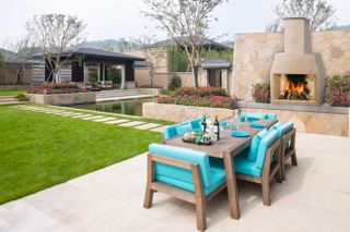 Wooden patio furniture ideas in front of an outdoor fireplace, with bright aquamarine seat cushions and dining plates.