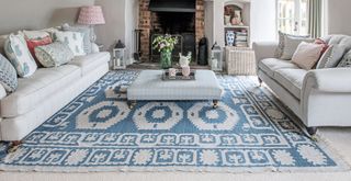 Neutral living room with large blue patterned rug over cream carpet in front of a working fireplace with two sofas facing each other with an ottoman coffee table