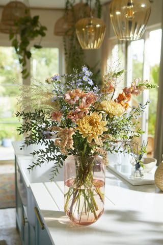 bunch of autumnal coloured flowers in a vase on a kitchen countertop, amber glass pendant lights, plants in the background
