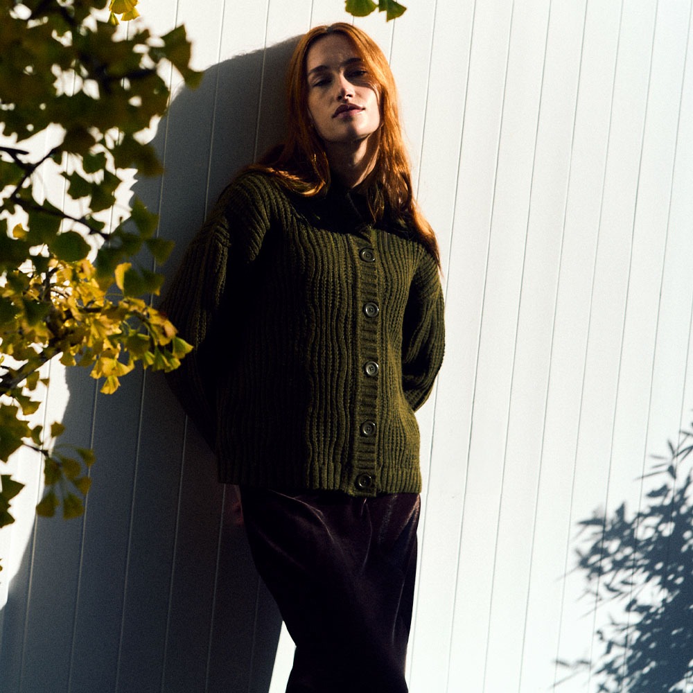 Model leaning against a garden fence wearing a green Finery London cardigan