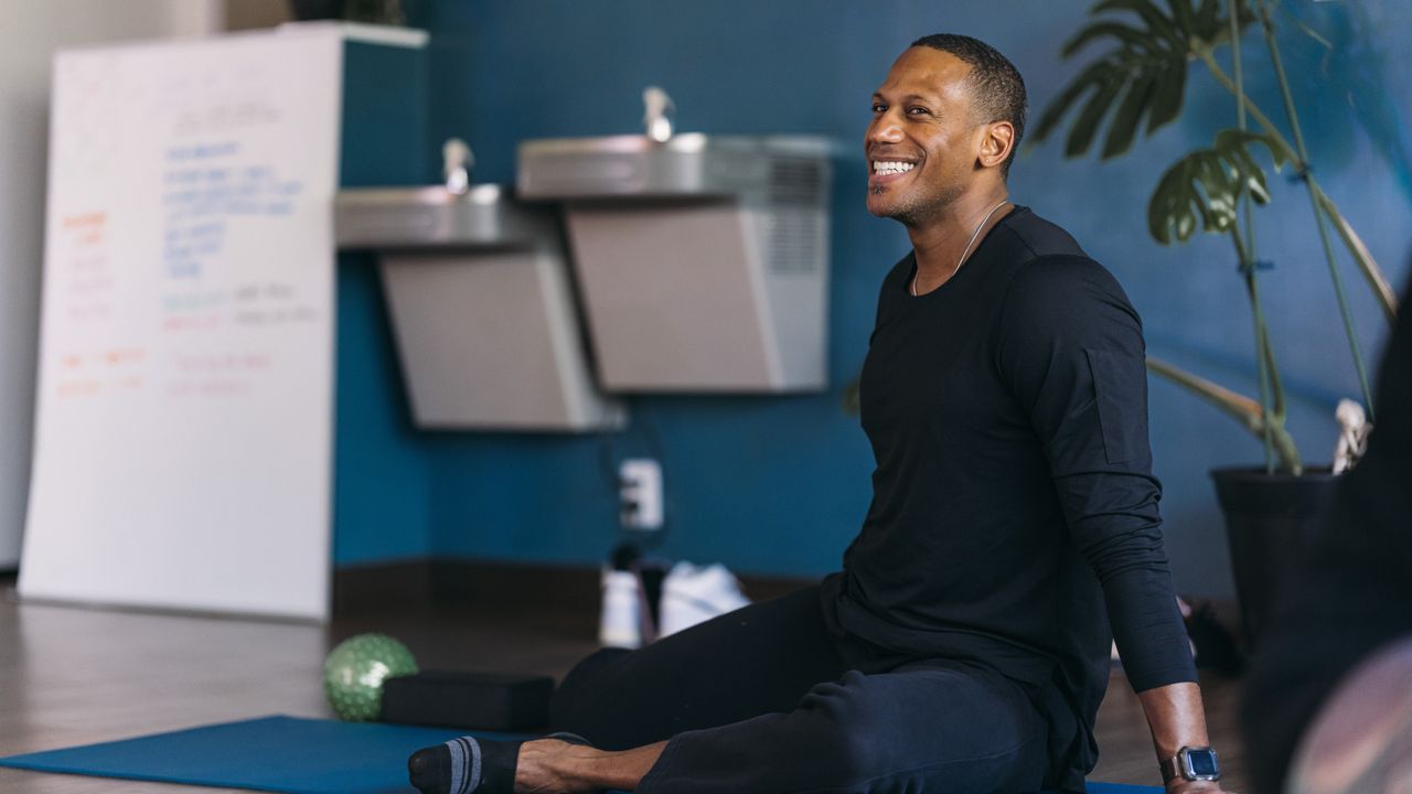 A man performs a 90/90 hip stretch in a yoga room. He is on the floor, with his legs bent; one is bent in front of him and the other is to the side. Behind him we see metal sinks and a plant.