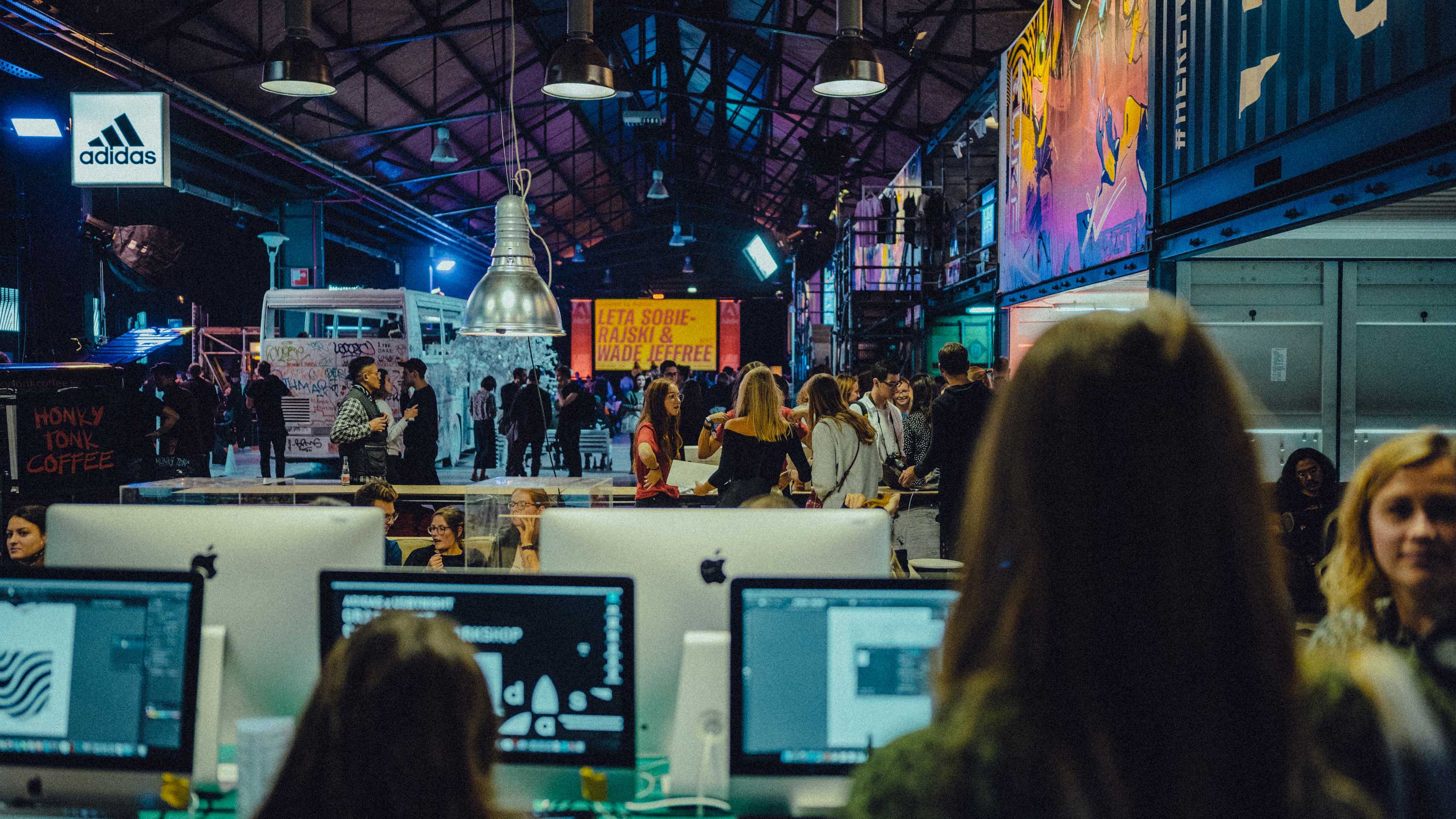A large warehouse with dangling light fixtures, open shipping containers line the right wall. In the foreground are multiple Mac computers being used by attendees. In the background people are walking around the instalments and market areas.