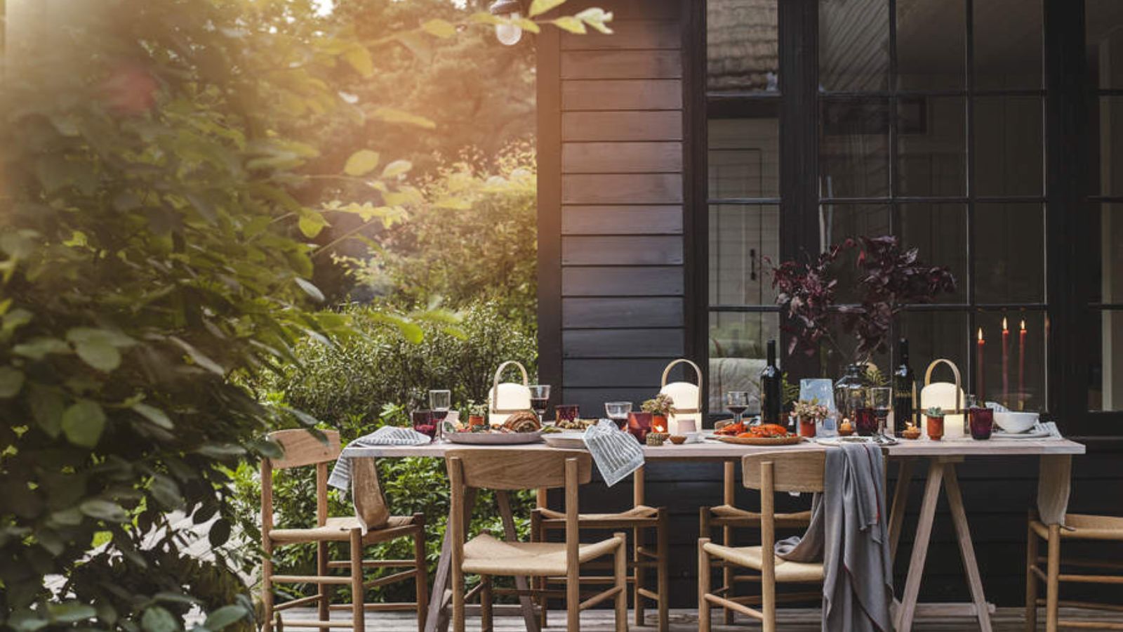 An outdoor dining table laid for lunch against dark wood walls and lush greenery. 