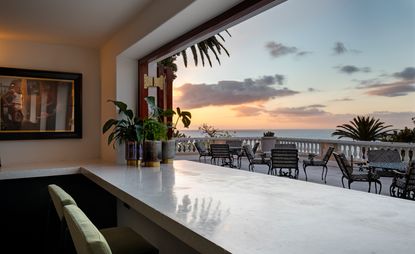 View of the terrace as the sun sets from inside Bar Roc at Ellerman House, South Africa. There is framed art on the wall, green plants in pots on a light coloured counter and pale green chairs