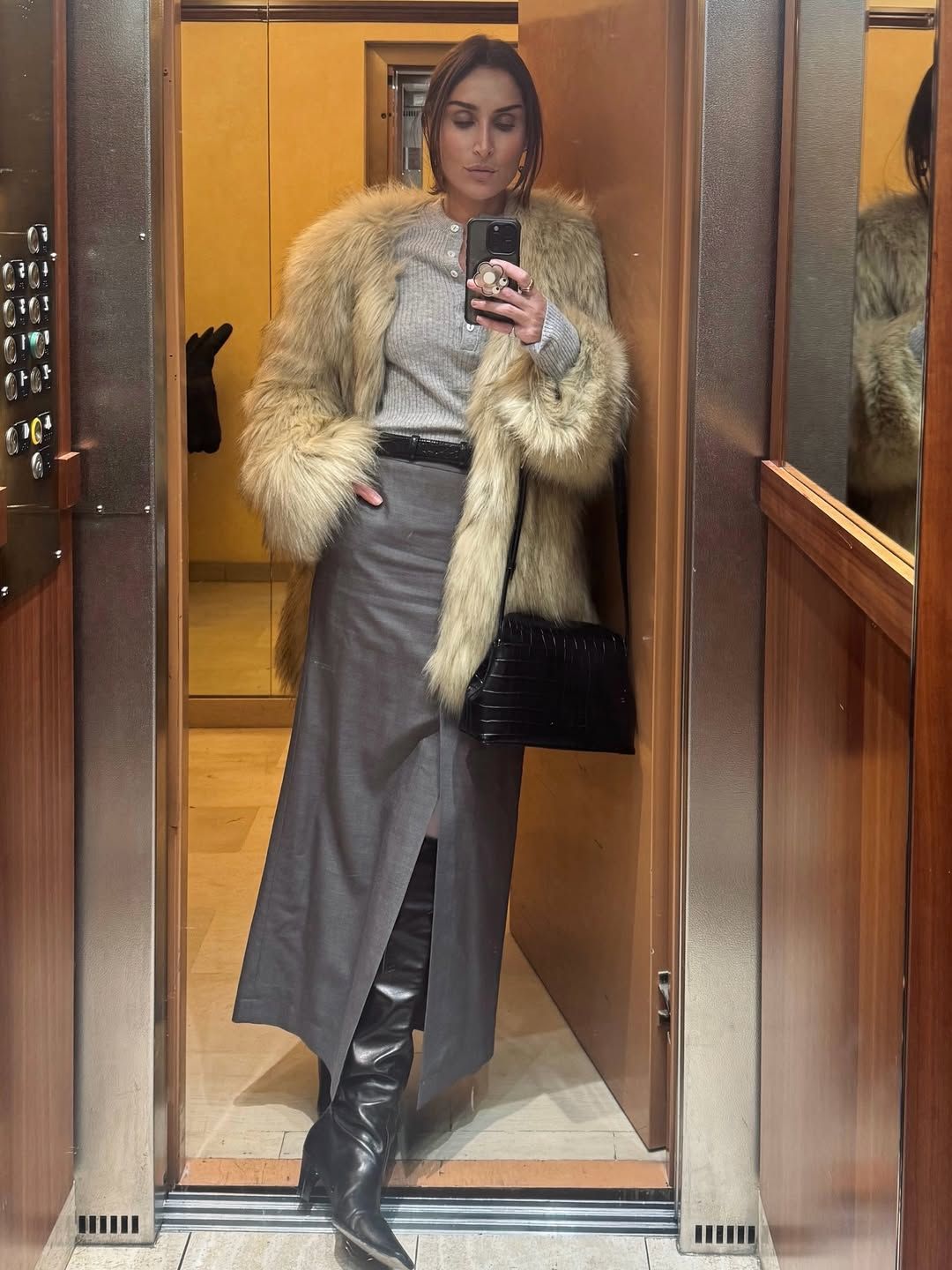 Géraldine wears a faux fur jacket with a henley shirt, gray skirt, and black leather boots.
