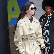 Mikey wears a trench coat, black pumps, black sunglasses, and a black ring-handle bag.