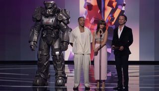 Three actors on stage with a person inside a suit of power armor