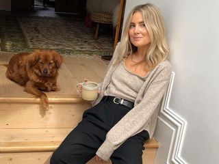 British influencer Lucy Williams poses with her dog on the steps of her London home wearing a neutral cardigan and matching top, black belt and black trousers.