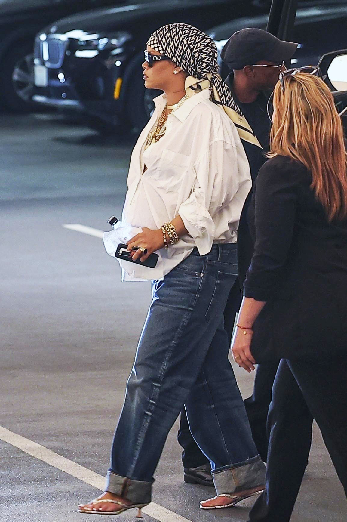 Rihanna wearing a silk scarf, black sunglasses, a white button down, blue jeans, and gold and pearl jewelry