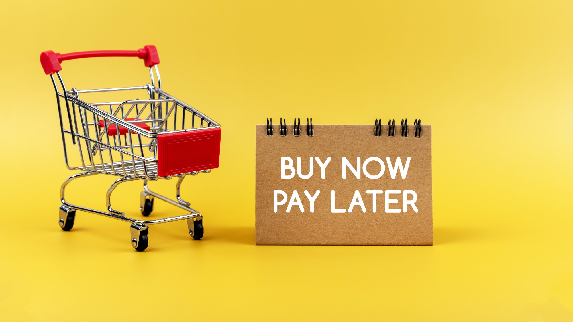 Miniature shopping cart next to a sign that reads "buy now pay later" on a yellow background 