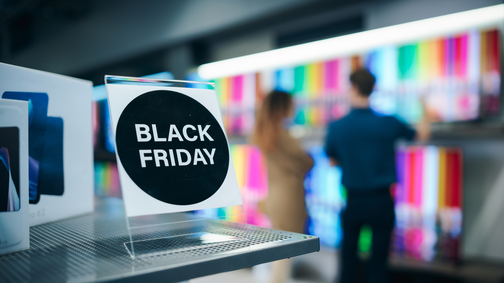 Two people stand in front of a retail wall featuring several TVs. In the foreground is a sign that reads: "BLACK FRIDAY".