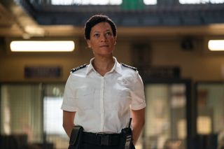 Leigh (Nina Sosanya) stands in the main atrium of the prison wing wearing her uniform, with her arms resting behind her back. She is looking slightly upward at something.