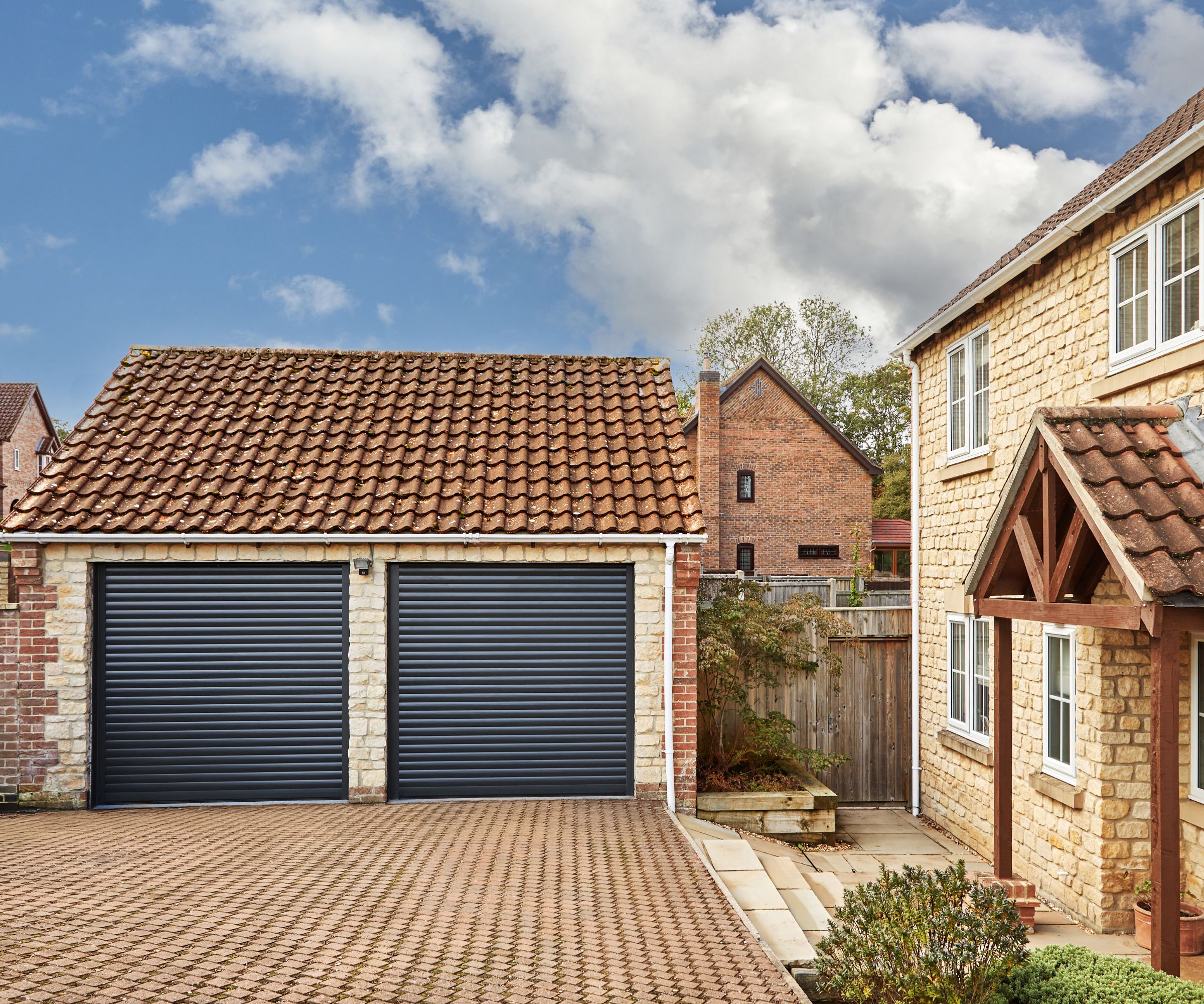 A charming countryside home with a warm-toned stone exterior, red tiled roof, and a wooden porch. The detached double garage features two black roller doors, set against a patterned brick driveway under a bright blue sky with scattered clouds.