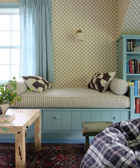 a daybed in a small New England guest cottage with ditsy floral wallpaper, blue painted daybed and storage with a striped cushion and bolster pillows