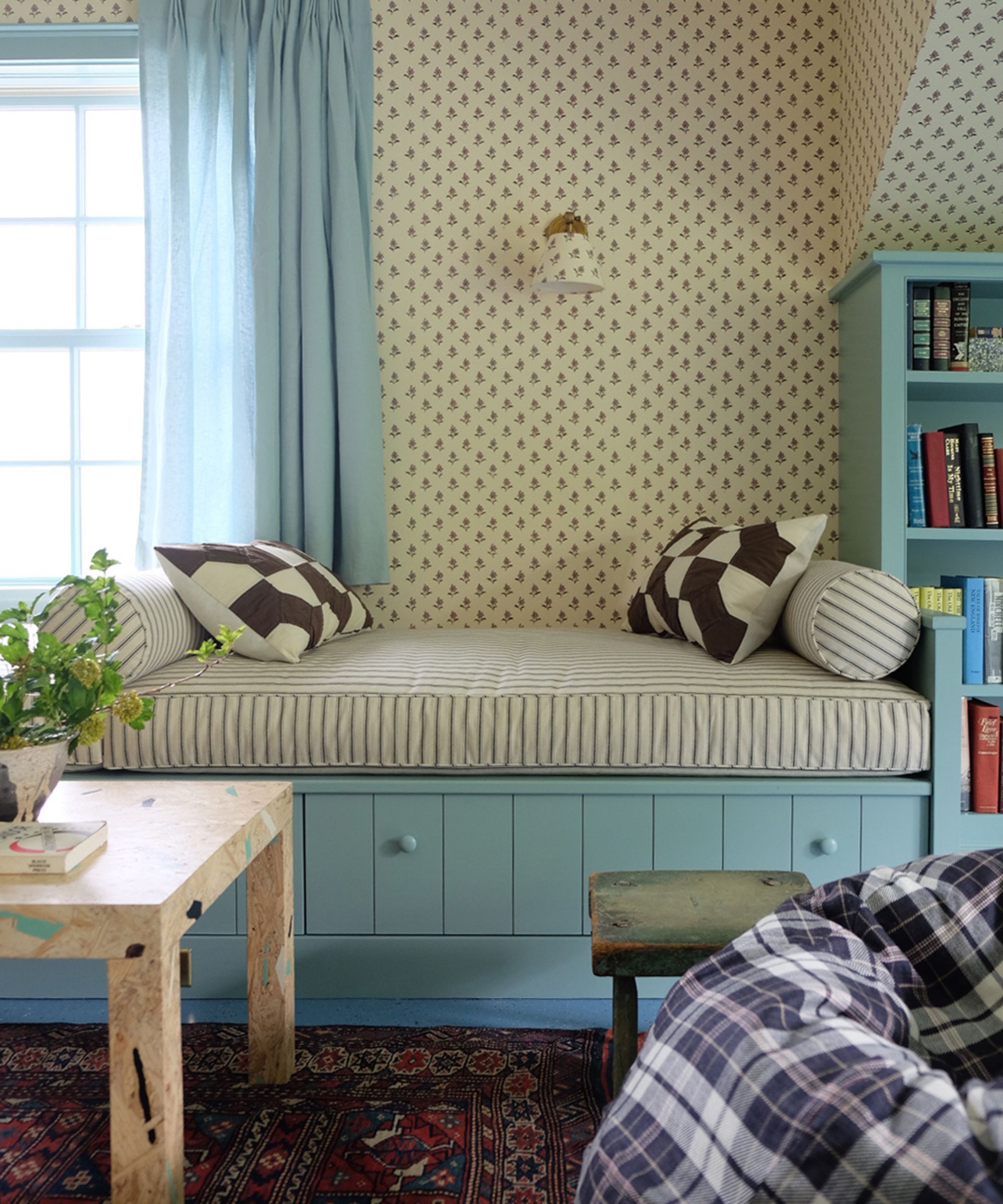 a daybed in a small New England guest cottage with ditsy floral wallpaper, blue painted daybed and storage with a striped cushion and bolster pillows