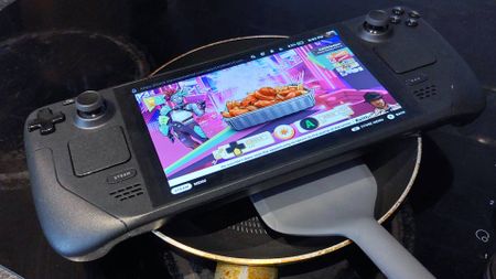 A surreal photo where a Steam Deck handheld gaming pc has been placed over the top of a much too small frying pan. Someone is attempting to flip the Steam Deck with a grey, silicone spatula.