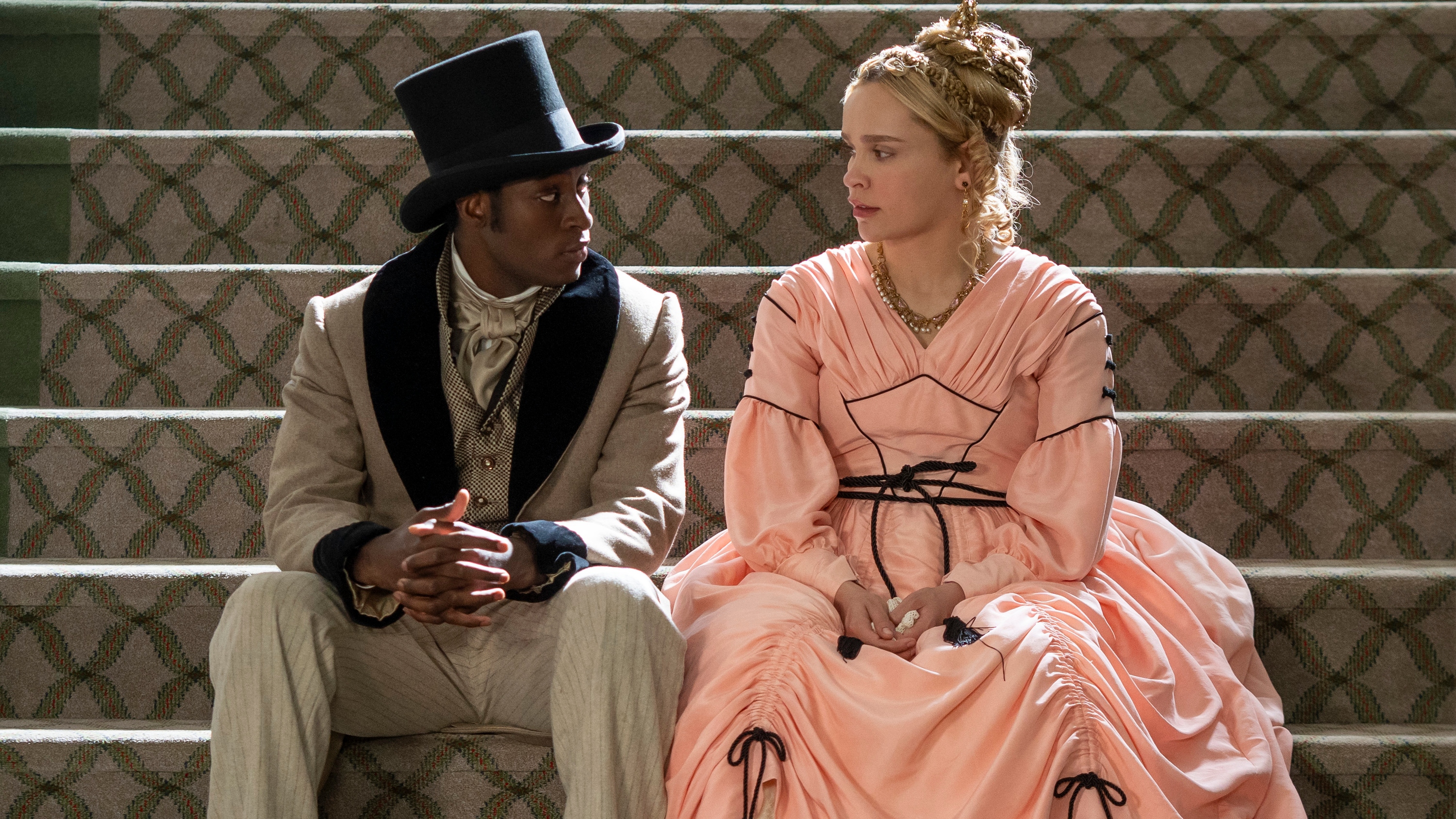Ernest Kingsley Junior and Iola Evans as Washington Black characters Wash and Tanna. Both are sitting on a lavish staircase, gazing at each other and dressed in upperclass 19th century clothing. Wash wears a light coloured suit and a top hat, while Tanna wears a pleated peach dress with black trim.